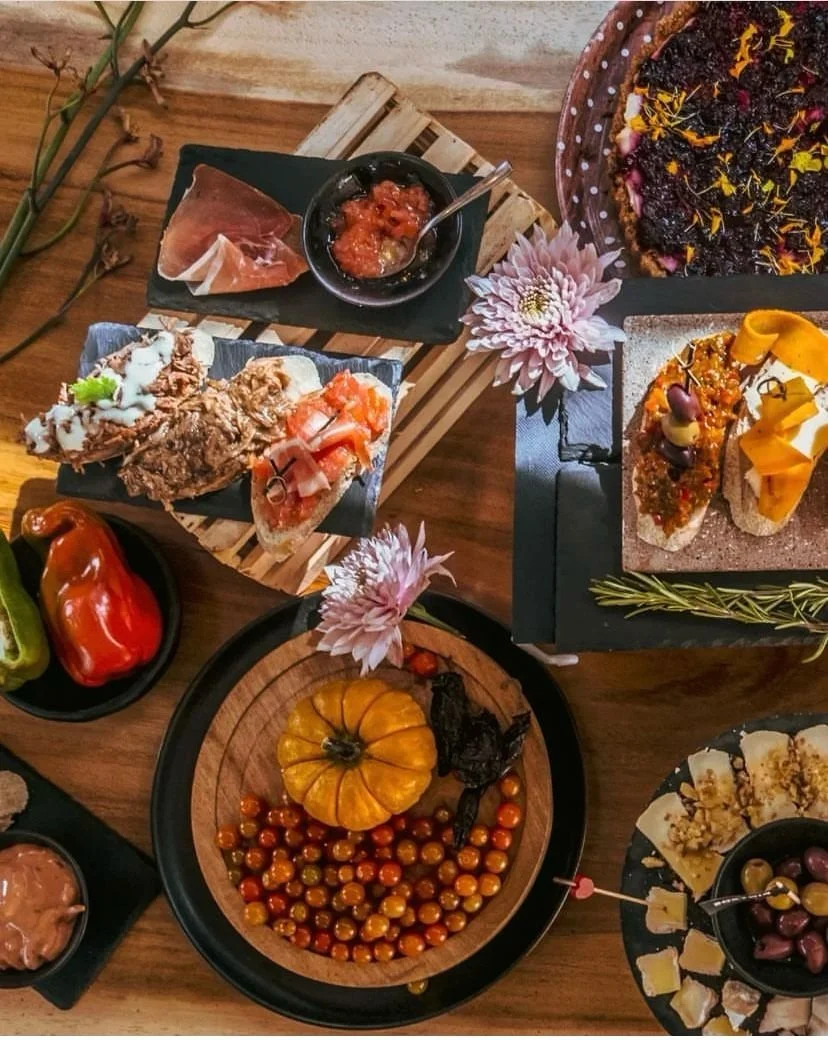 Overhead view of an assorted charcuterie spread featuring meats, cheese, olives, fruit, and small decorative pumpkins on a wooden table with decorative flowers.