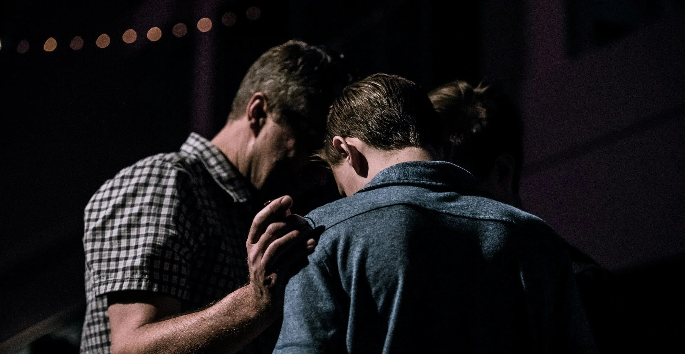 Three individuals in dark clothing praying or having a moment of silence with their heads bowed and eyes closed in a dimly lit setting.