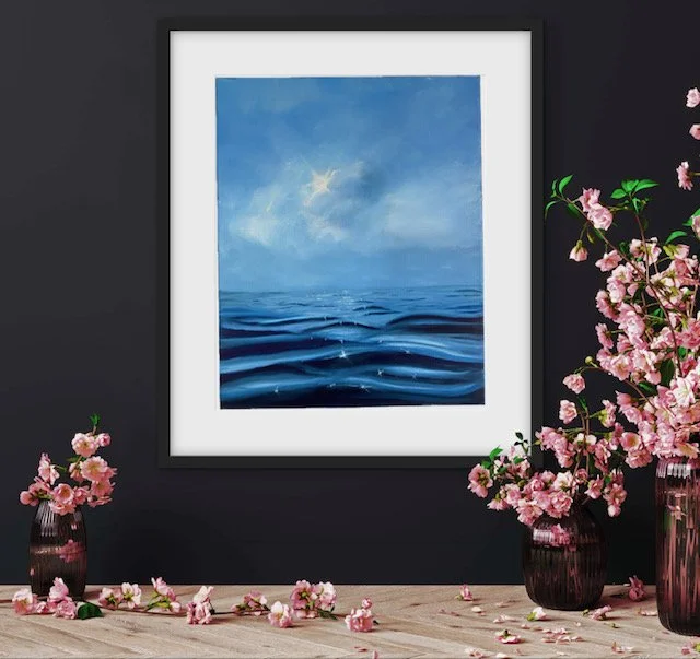 Painting of blue sky and ocean waves hanging on a dark wall, flanked by pink cherry blossom vases with flowers and petals scattered on a wooden surface.