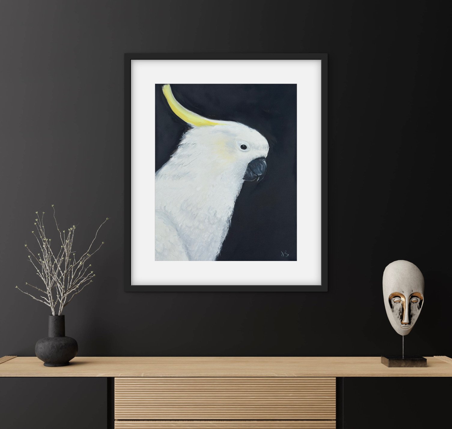 Wall art of a white cockatoo with a yellow crest, framed and hung on a dark wall, with a vase holding a bare branch, and a decorative mask on a stand on a light wood surface.
