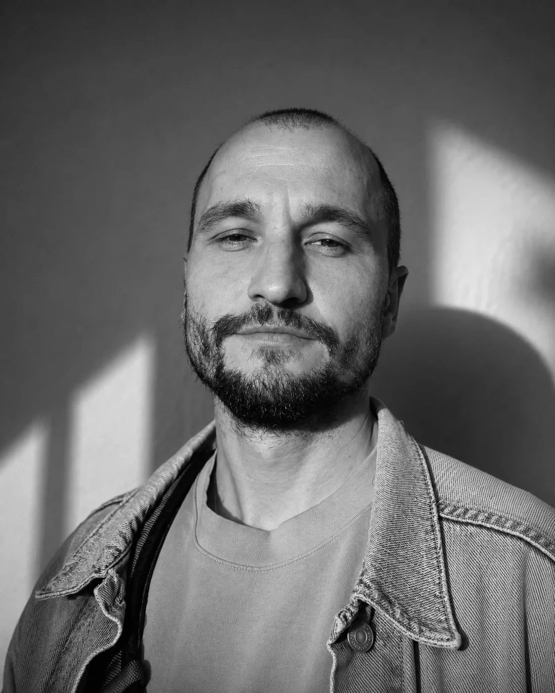 Black and white portrait of a man with a beard, short hair, wearing a denim jacket and t-shirt, standing against a plain wall with light and shadow patterns.