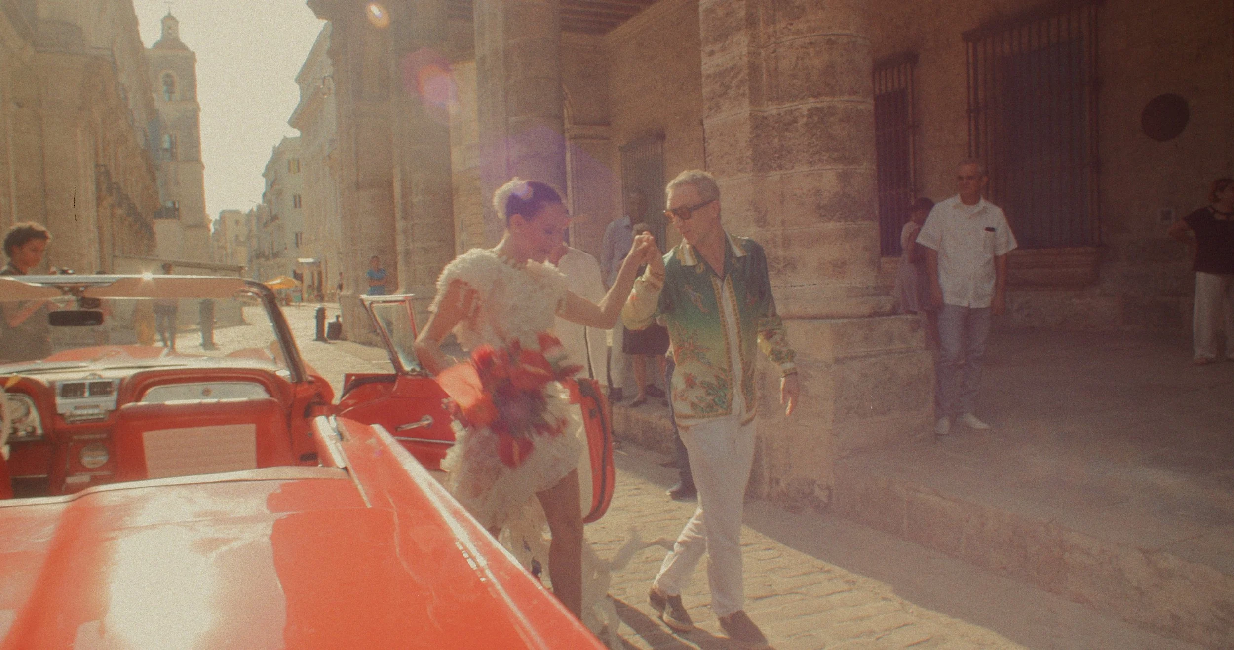 People walking on a street with a vintage red convertible car in the foreground, sun shining brightly, and old stone buildings in the background. Wedding in Havana, Cuba.