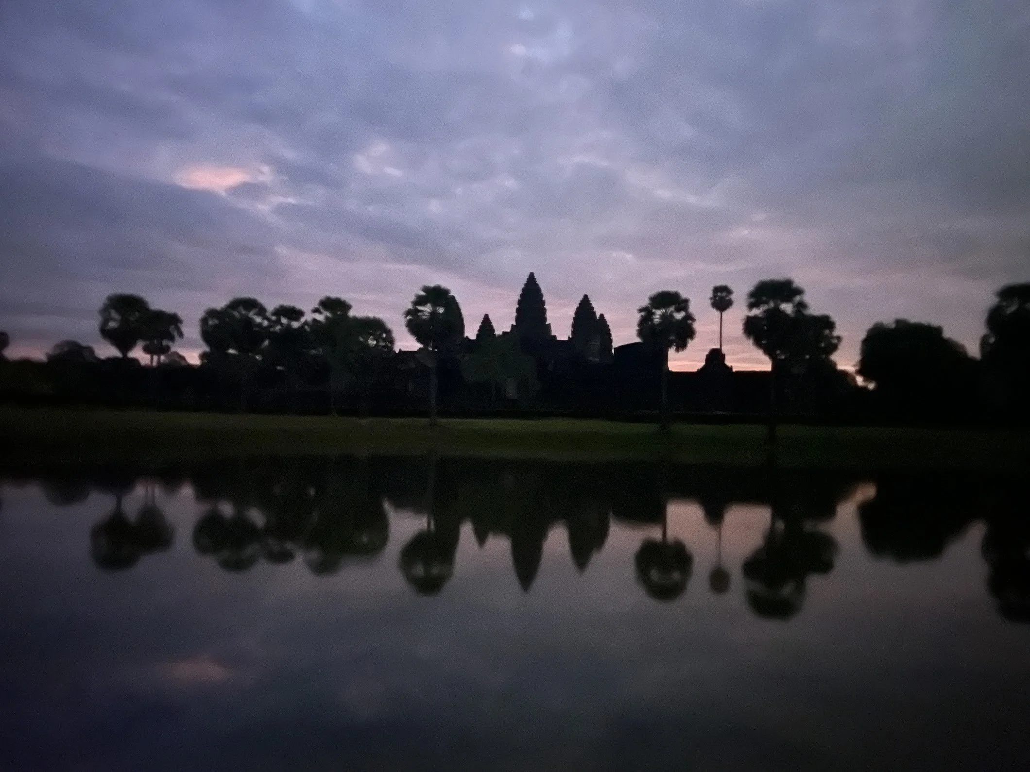 Silhouette of Angkor Wat temple at sunrise or sunset with cloudy sky, mirrored over a body of water, with trees surrounding the area.