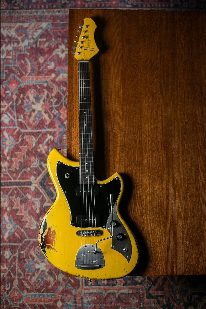 Yellow electric guitar with black pickguard, partially worn paint, placed on a wooden surface, with a patterned rug underneath.