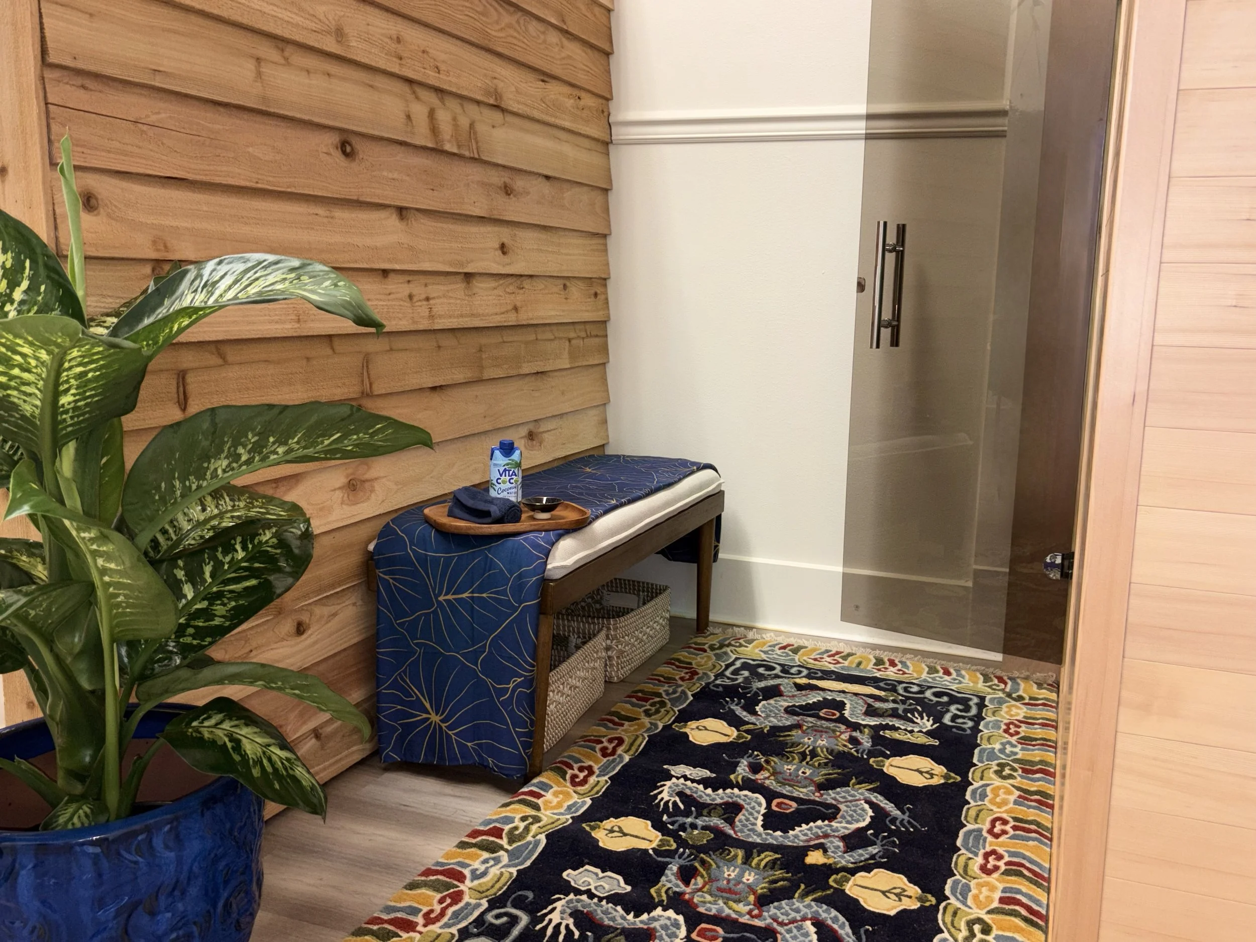 Interior view of a Hilo Sauna with a wooden accent wall, a patterned rug on the floor, a potted plant in the foreground, a bench with a blue cover, a basket underneath, a tray with a water bottle and small dish, and a glass door on the right.