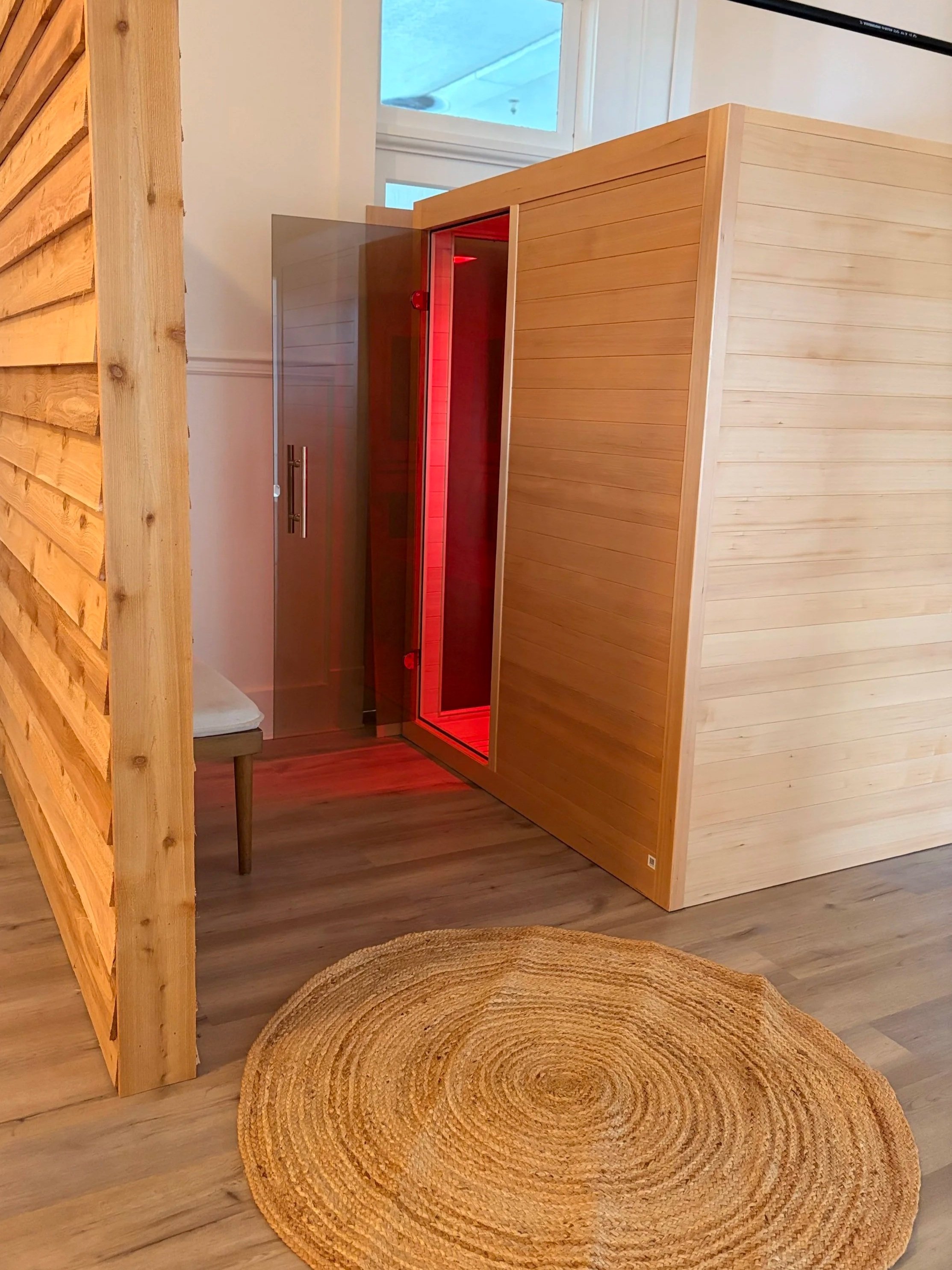 A Hilo Infrared Sauna room at Yoga Centered with wooden walls and a glass door with red lighting inside. There is a small stool next to the door, a window above with a view outside, a wooden partition, and a circular woven rug on the floor.