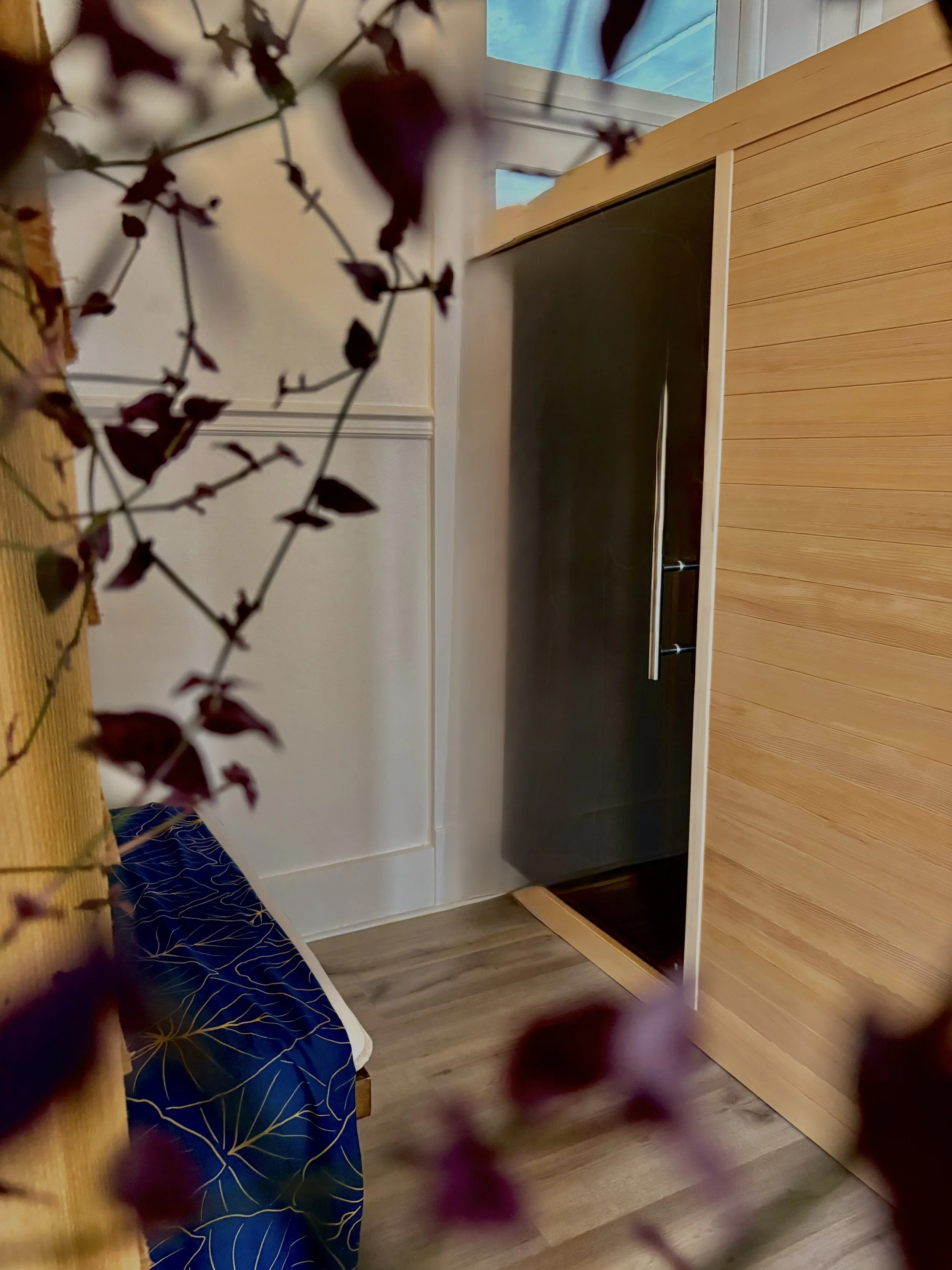 Interior of a room with wooden floor, white wall, and sliding door leading to the Yoga Centered Hilo Infrared Sauna. There is a bench with blue and yellow patterned bedspread on the left side.