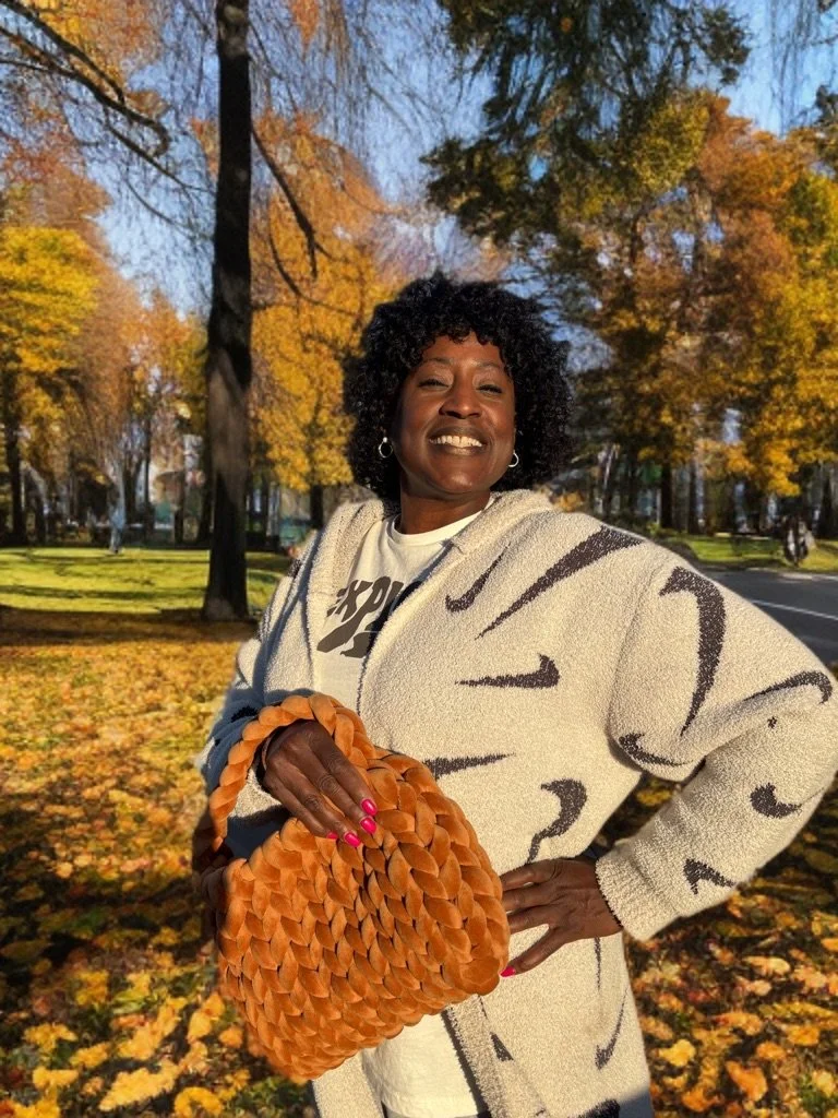 A woman smiling in a park with fall leaves on the ground, wearing a patterned coat and holding an orange woven bag.