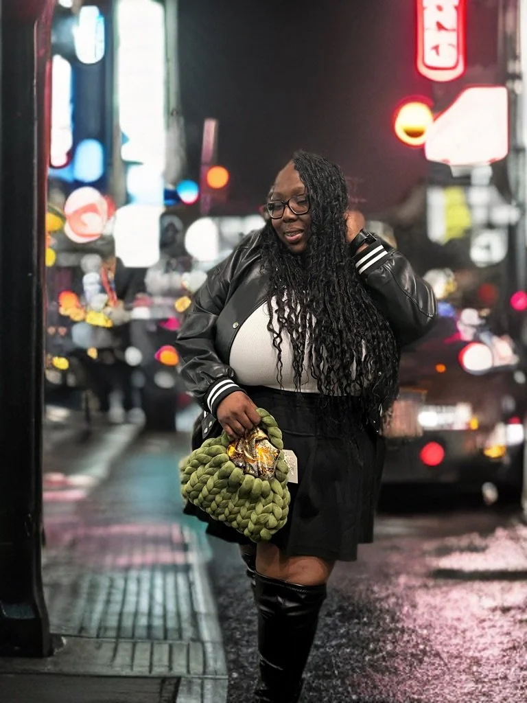 Woman in urban nightlife setting, wearing a leather jacket and glasses, holding a green handbag. Bright, colorful neon signs in the background.