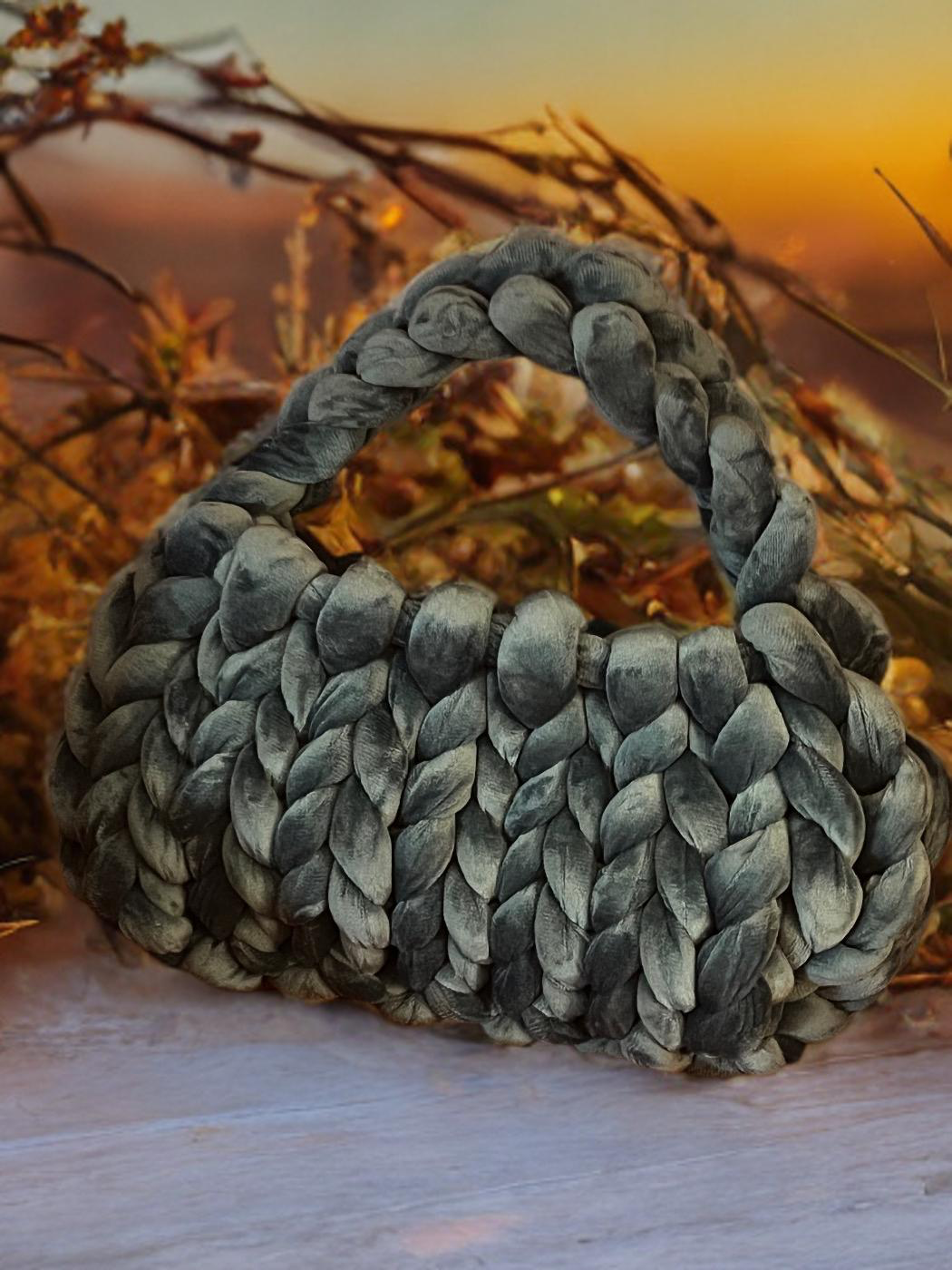A gray, knitted handbag with a handle, styled in a chunky braided pattern, placed outdoors on a wooden surface with dry plants and a sunset in the background.