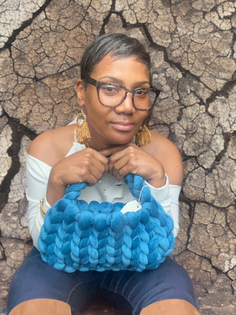 Person holding a blue knitted bag in front of a cracked earth background, wearing glasses and tassel earrings.