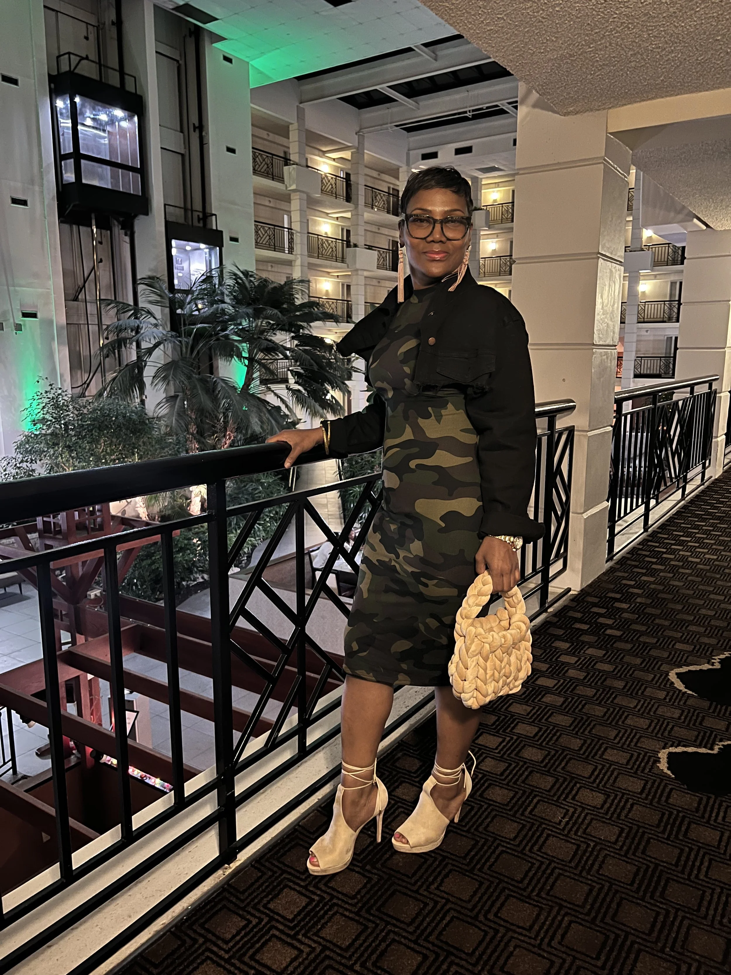 Woman posing on hotel balcony in camo dress and holding a woven handbag, with elevators and palm trees in the background.