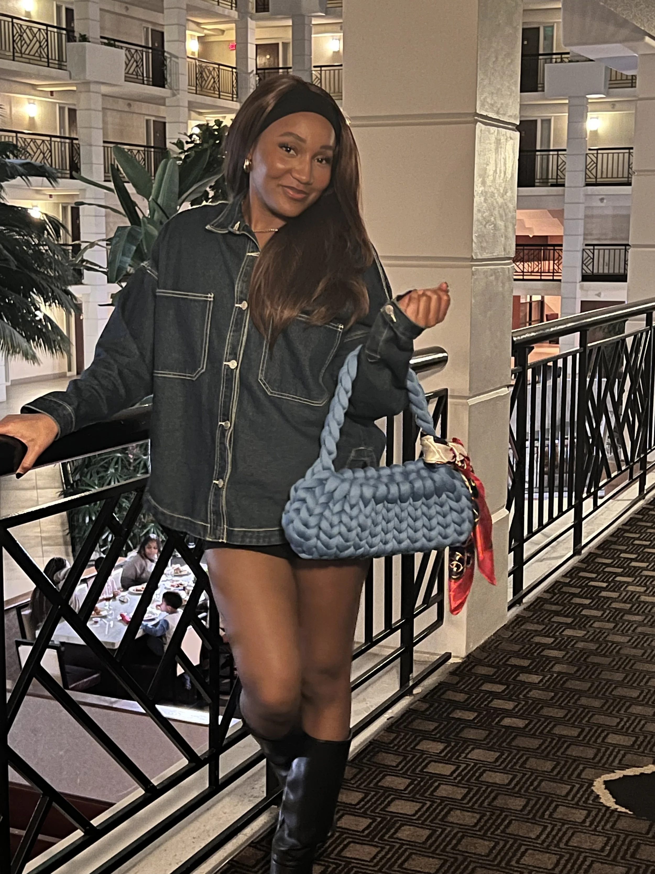 Woman posing in a hotel atrium, wearing a denim jacket, headband, and holding a blue woven handbag. Indoor plants and dining area visible in the background.