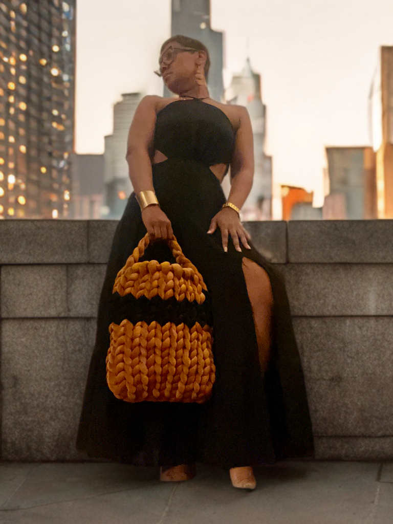 Woman in black dress with a large woven bag standing against a city backdrop at dusk.