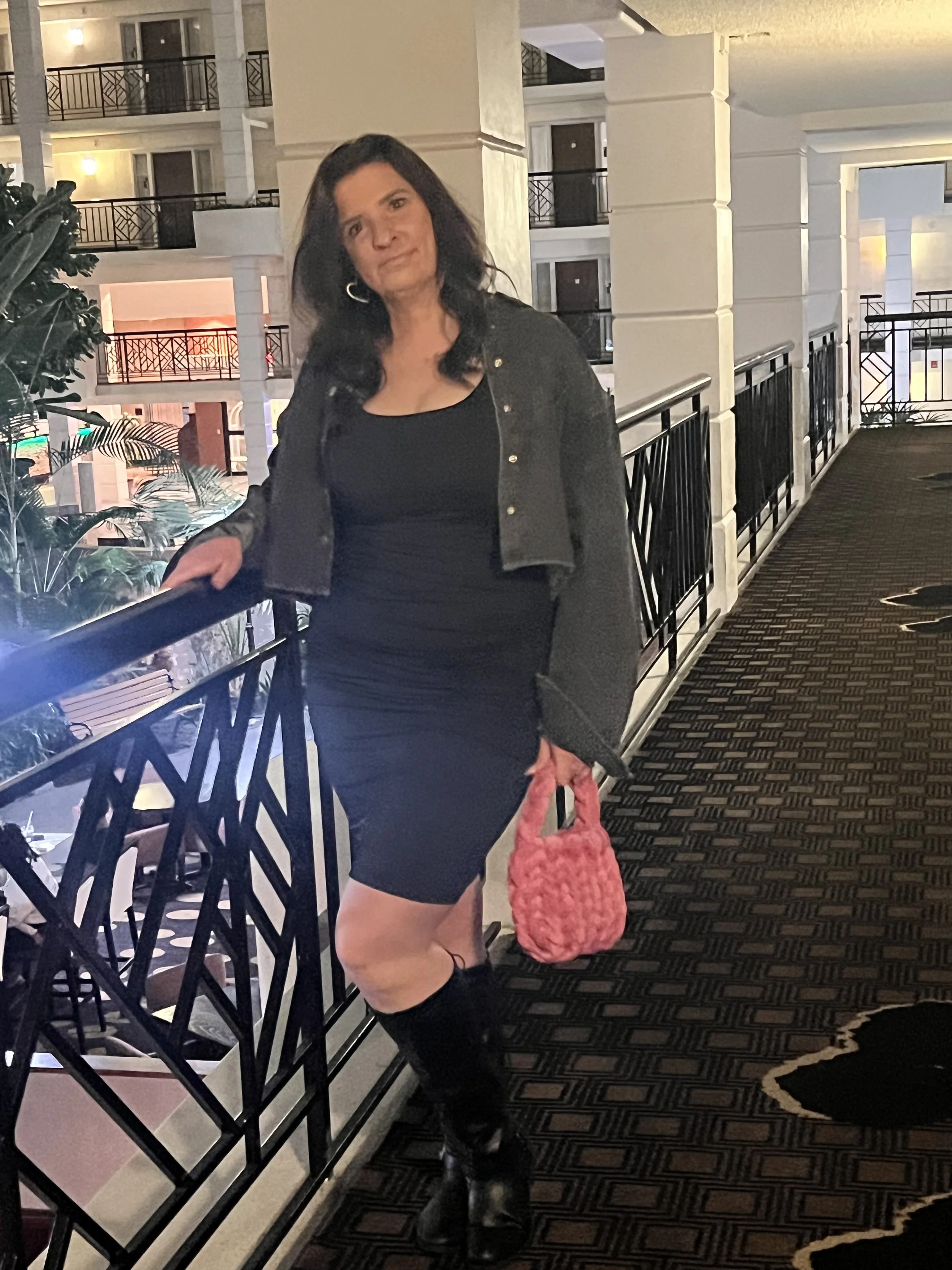 Woman standing on a hotel walkway, wearing a black dress, a gray jacket, and holding a pink bag.