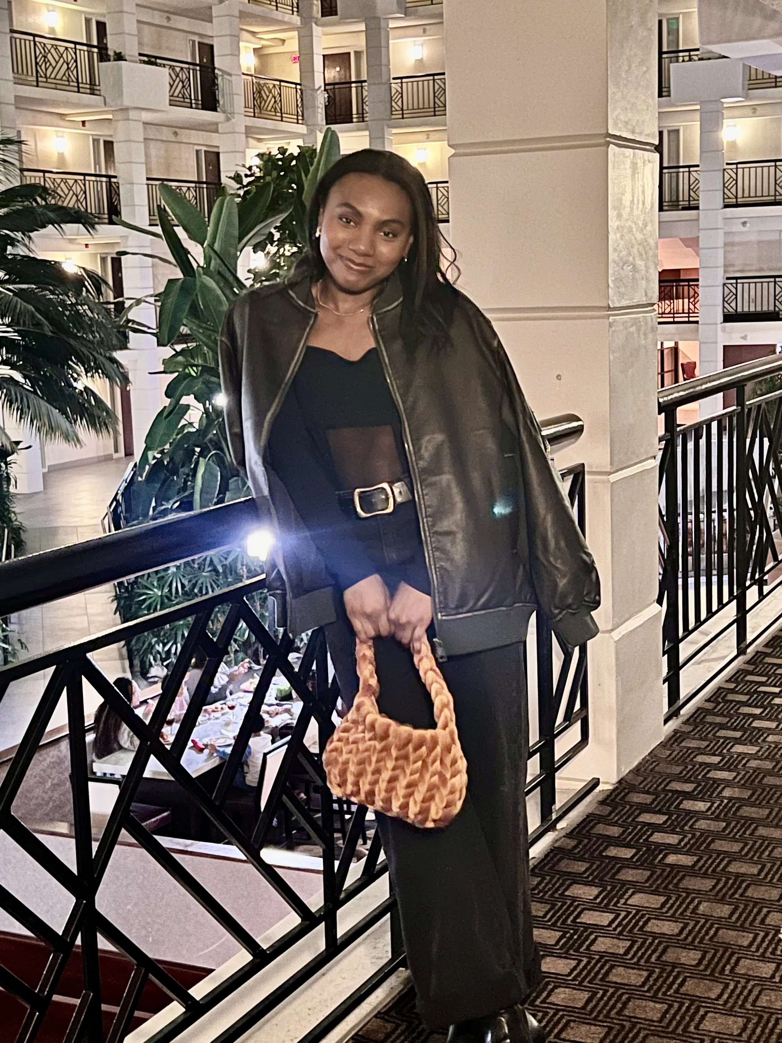 A person wearing a black outfit with a leather jacket stands in a hotel atrium, holding a textured handbag. The background features balconies, plants, and a dining area below.