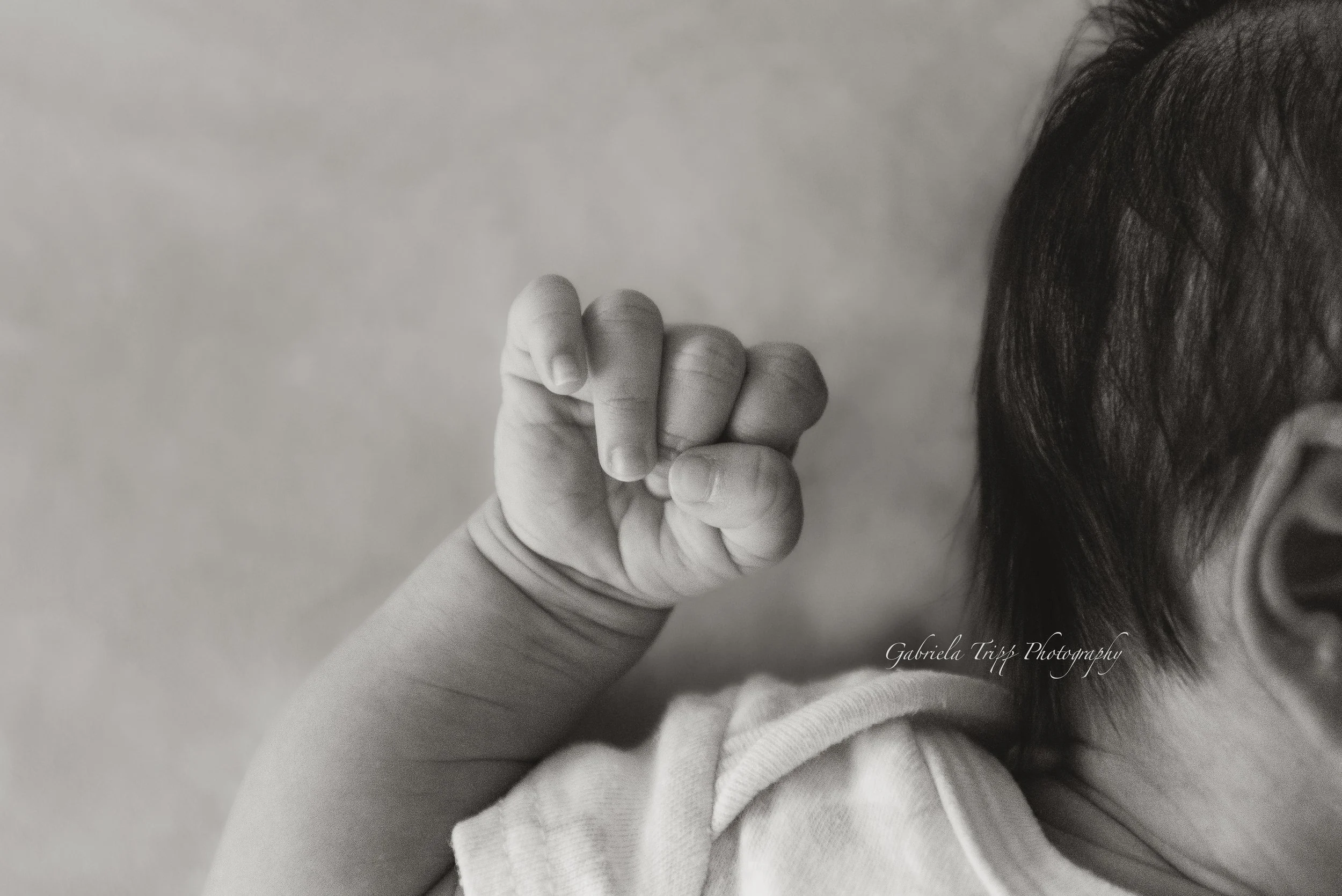 A newborn photograph with close up on baby's hand, with part of the child's face and hair visible in the background.