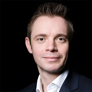 A man with short dark hair and fair skin, wearing a dark blazer and white shirt, smiling slightly against a black background.