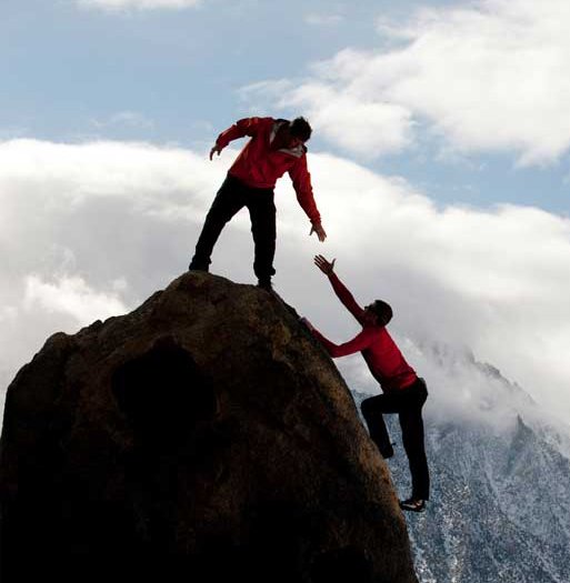 Two people on a large rock, one reaching down to help the other climb up, with a mountain and cloudy sky in the background.