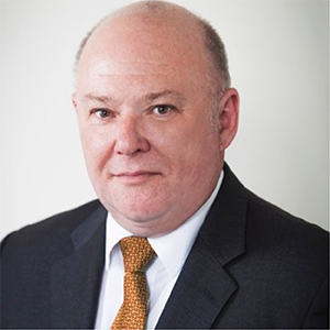 Professional headshot of a middle-aged man wearing a dark suit, white shirt, and a gold tie, with a neutral background.