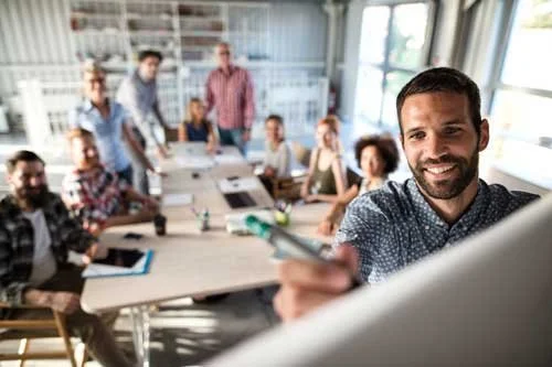Man taking writing on whiteboard in a bright conference room with a group of people sitting and standing around a large table representing supply chain risk training
