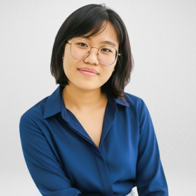 A young woman with glasses and short black hair, smiling and resting her chin on her hand in a warmly lit room.