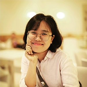 A young woman with glasses and short black hair, smiling and resting her chin on her hand in a warmly lit room.