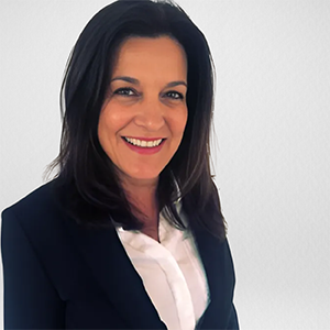 A woman with dark hair smiling, wearing a white shirt and a dark blazer against a plain background.