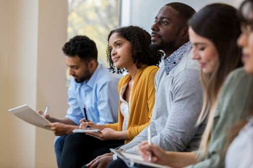 Group of diverse people attending a seminar or workshop, sitting in a row with notebooks and pens representing capability assessment training