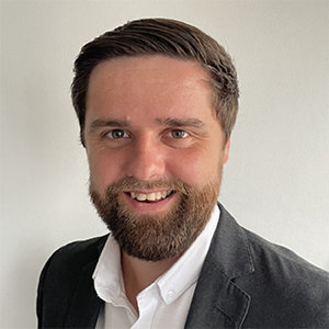A smiling man with short brown hair and a beard, wearing a white shirt and dark blazer, against a plain light background.