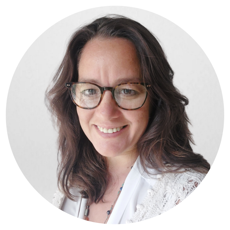 A woman with dark wavy hair wearing glasses, smiling, and dressed in a white top and lace cardigan.