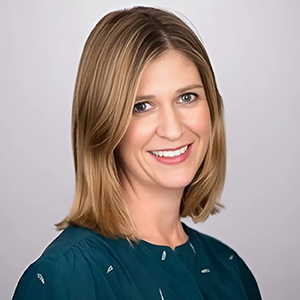 A woman with shoulder-length blonde hair smiling, wearing a teal shirt with white patterns, against a light background.