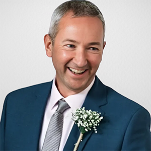 A man smiling, dressed in a blue suit and tie, with a white shirt and a boutonniere on his lapel, against a plain light background.