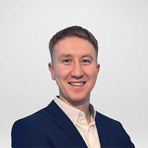 A young man with short brown hair smiling in front of a plain white background, wearing a navy blazer and white shirt.