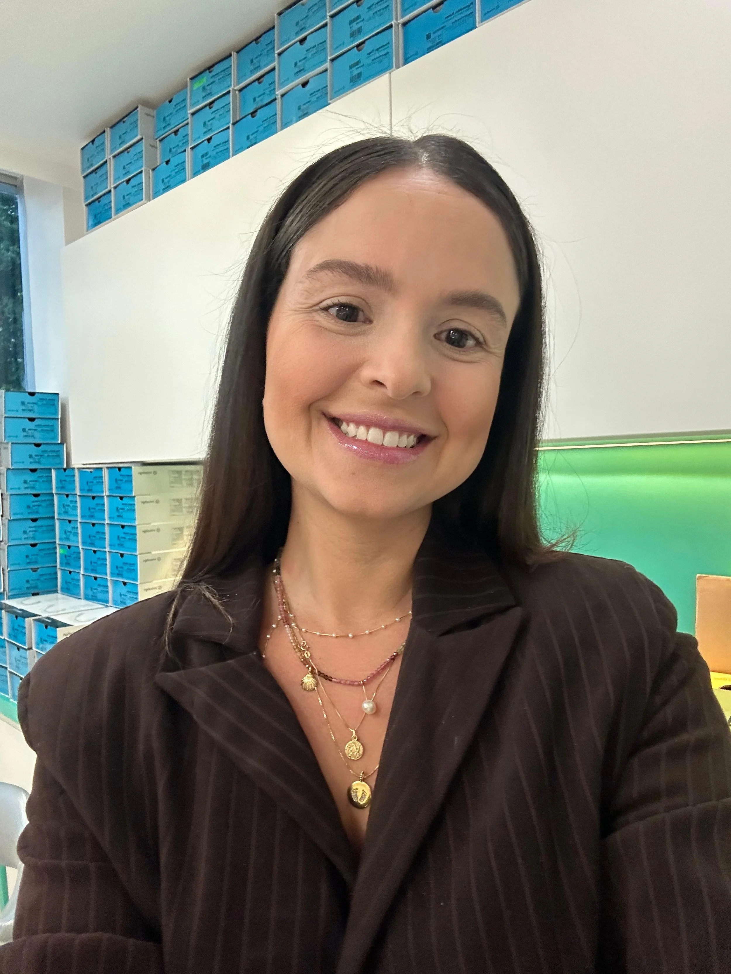 Mulher sorrindo em uma sala com prateleiras cheias de caixas azuis ao fundo.