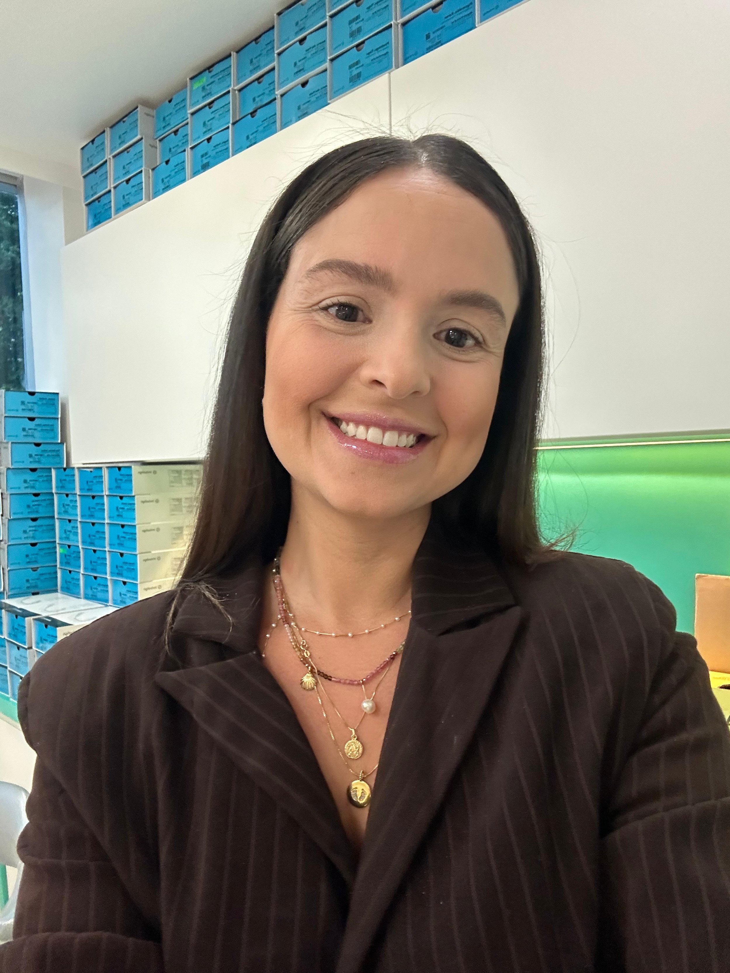 Mulher sorridente em uma sala com caixas azuis empilhadas nas prateleiras ao fundo.