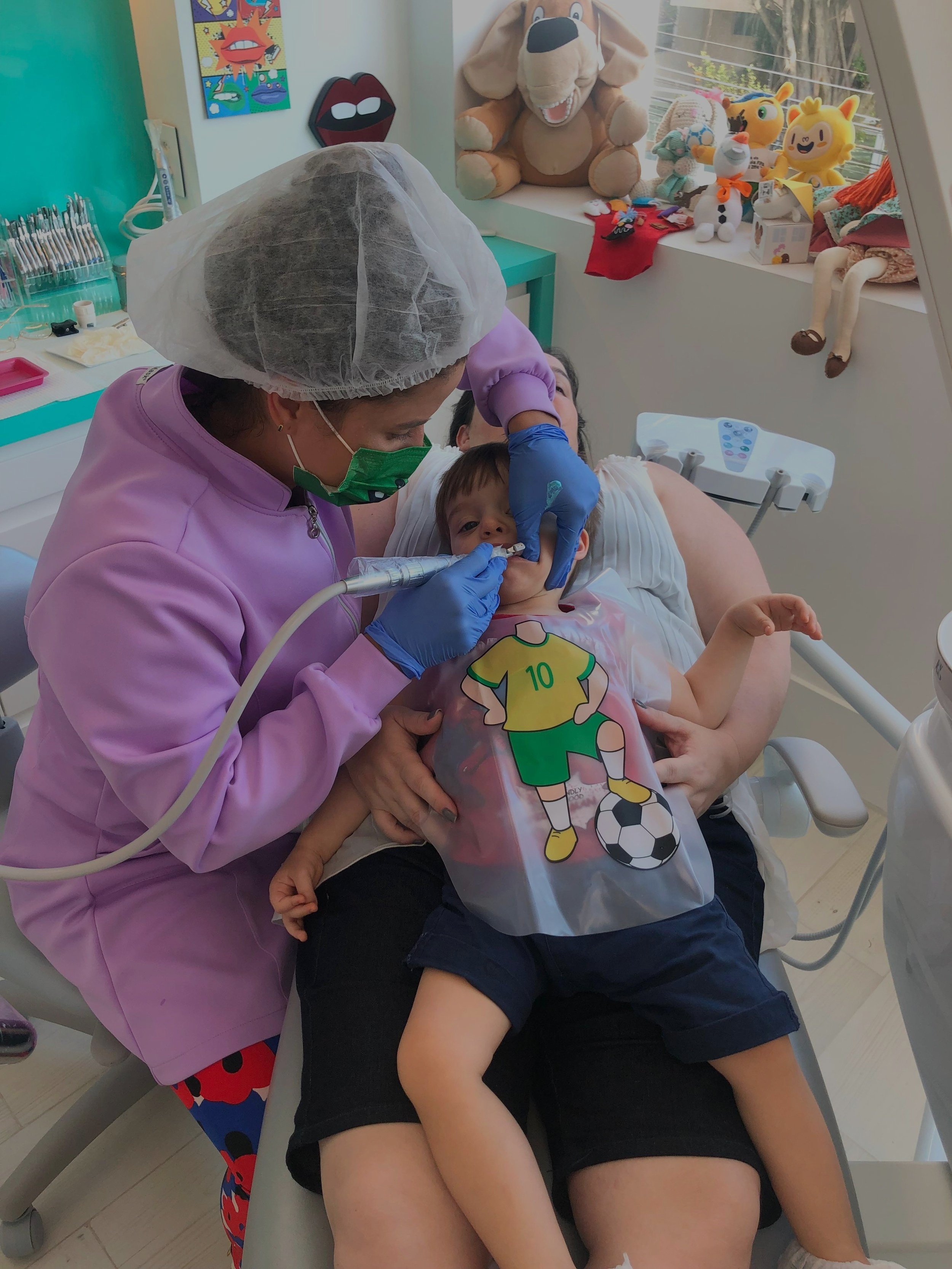 Dentista atendendo criança em consultório infantil colorido, com brinquedos e decoração lúdica ao fundo. Criança está no colo de um adulto, vestindo um avental com tema de futebol.