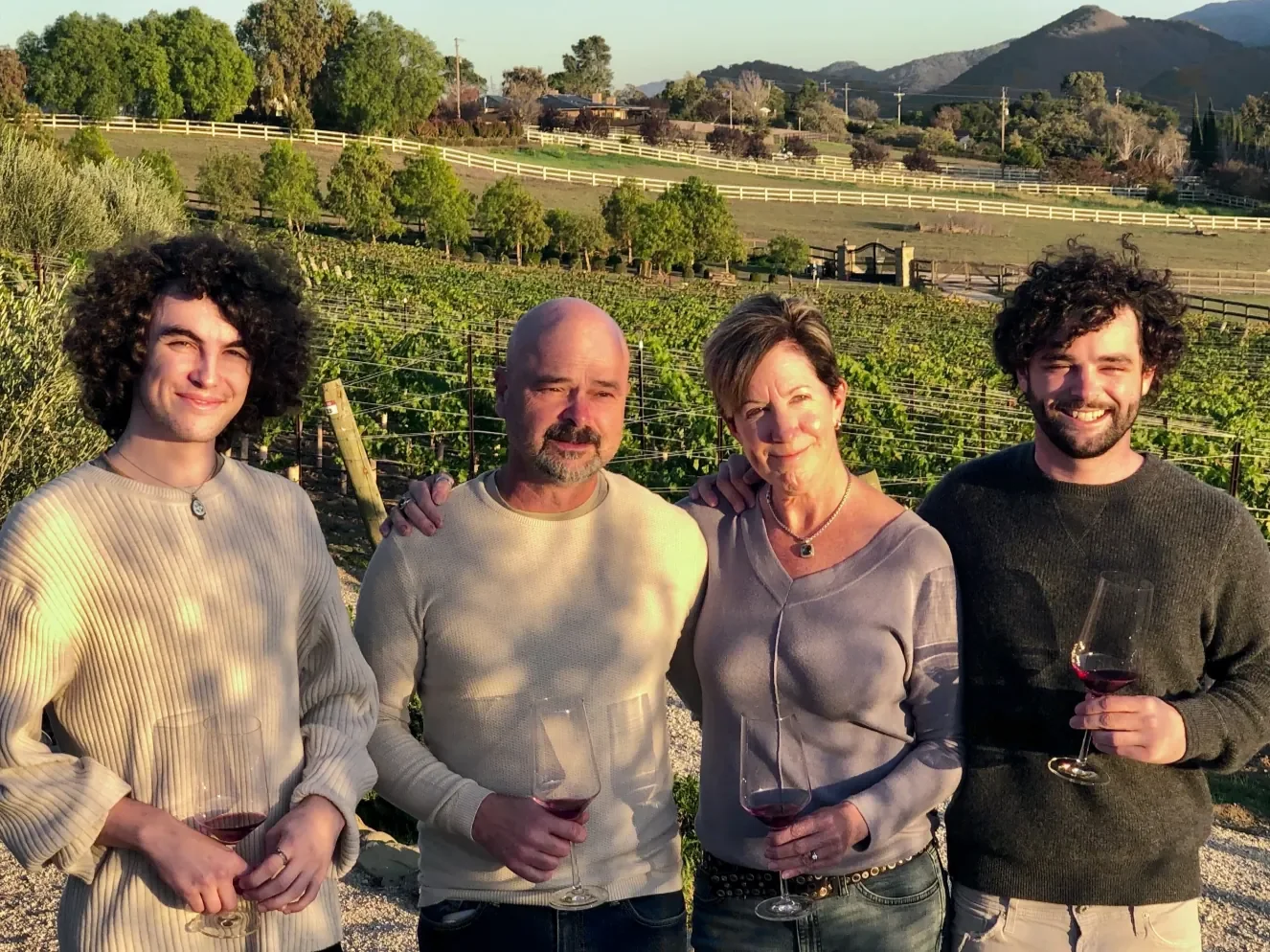 INÉSUEÑO Exquisite Bordeaux-style Blends Santa Ynez Valley Los Olivos District AVA - founders Julio and Colleen Estrada with sons Aidan and Julio Dylan standing together enjoying a glass of wine with the vineyard behind them.