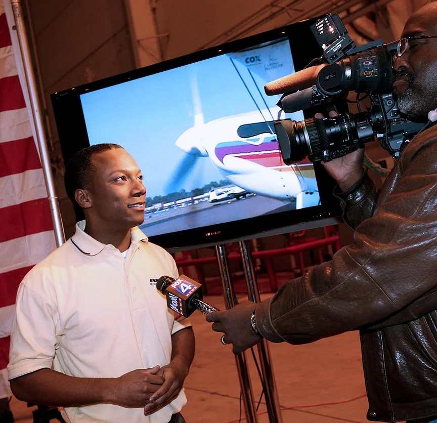 Man being interviewed by a news crew beside a television displaying an airplane image.
