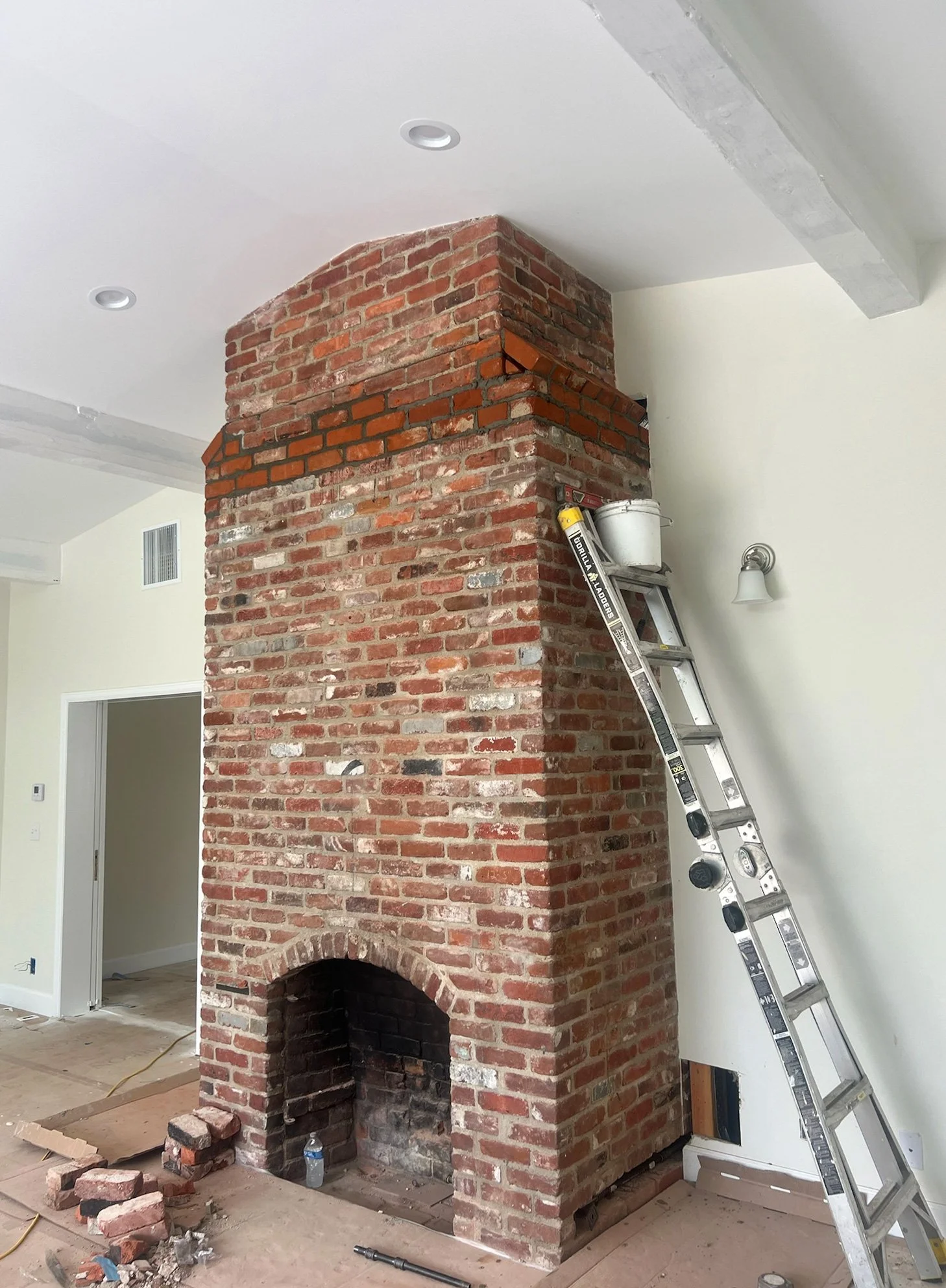 Brick fireplace construction in progress with a ladder leaning against it, construction tools, and materials inside a room.