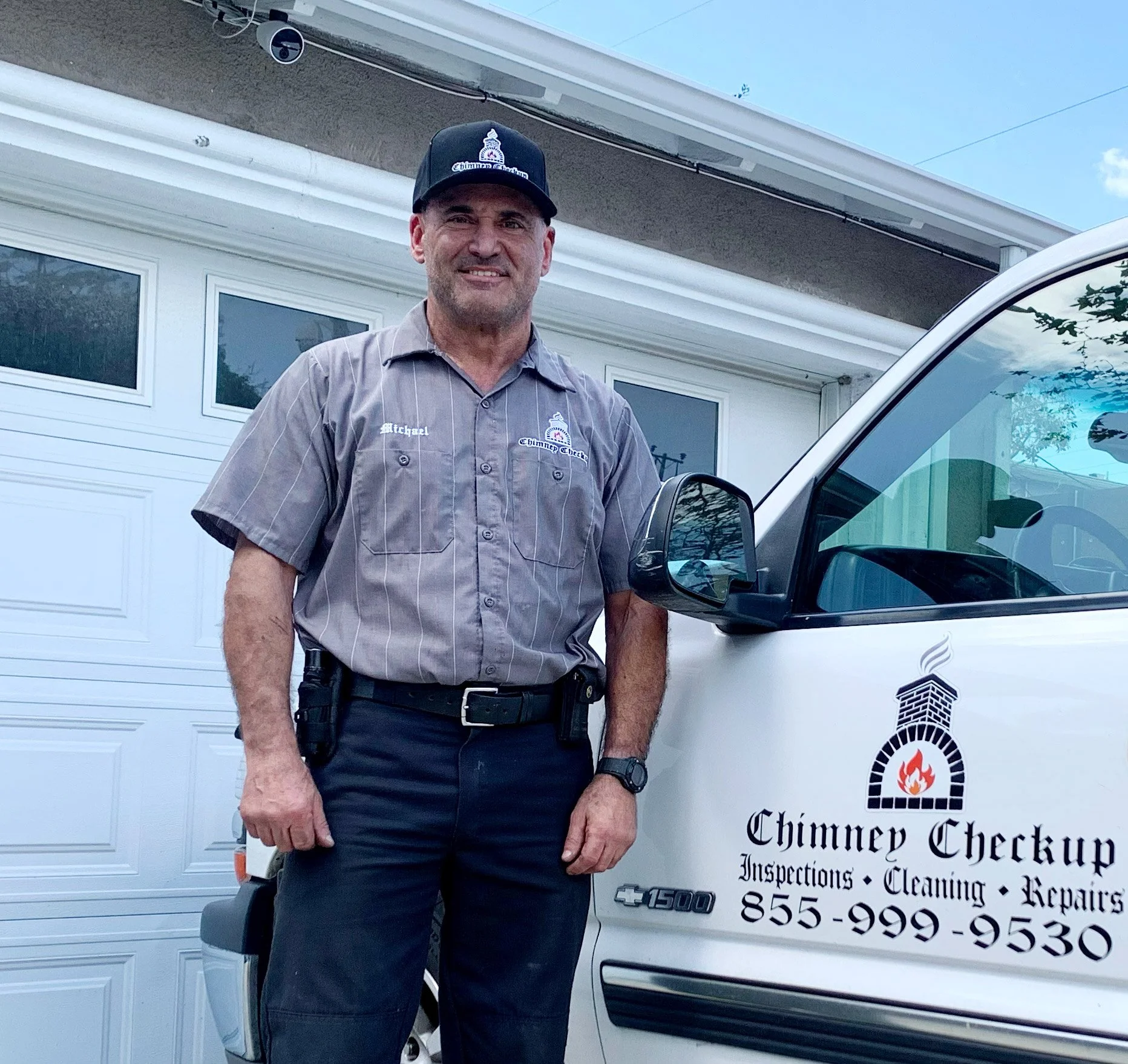 A chimney inspection and repair professional standing next to a branded vehicle in front of a house garage. The man is wearing a uniform shirt with the name 'Michael' and company logo, smartwatch, and a baseball cap with a chimney logo.