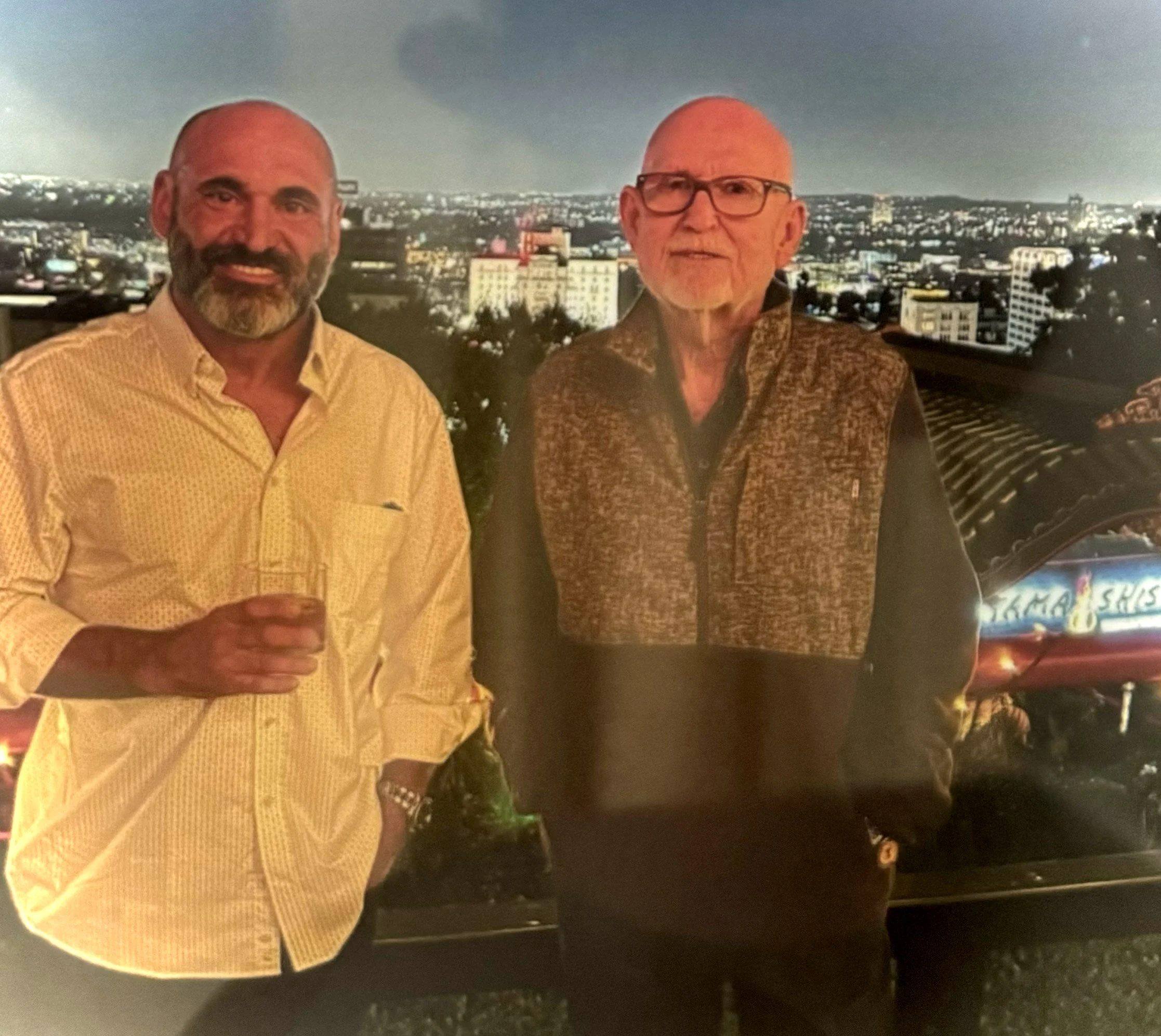 Two men standing with a city skyline at night in the background, one holding a drink.