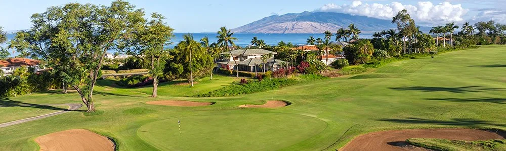 A scenic golf course with sand traps, green grass, trees, and residential houses, with a mountain and ocean in the background under a partly cloudy sky.