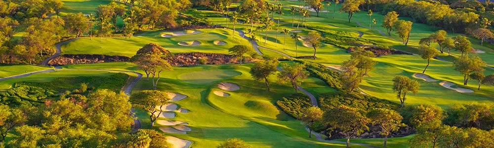 Aerial view of a golf course with lush green fairways, sand traps, trees, and rolling terrain.