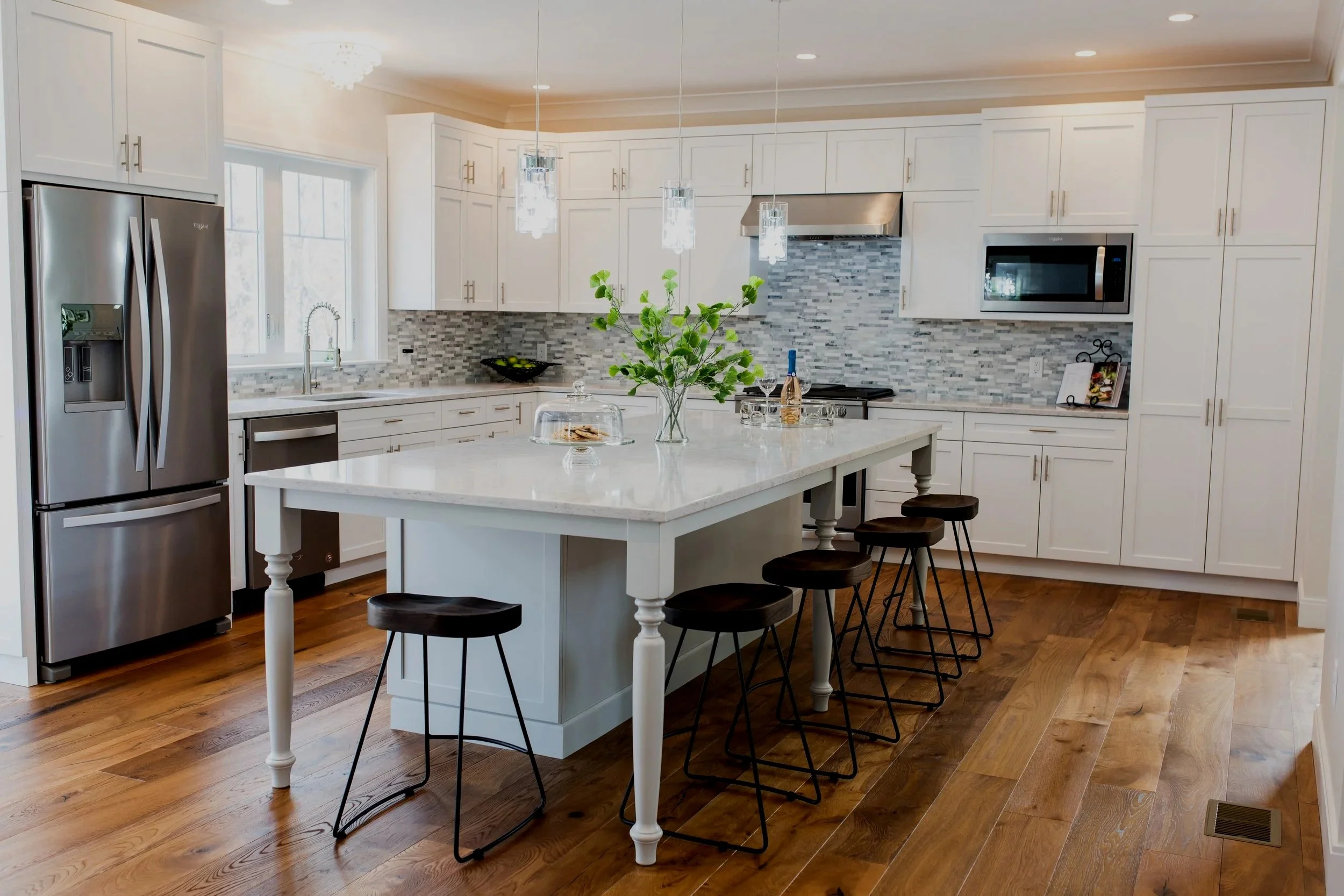 Modern kitchen with white cabinets, stainless steel refrigerator, marble island with bar stools, tiled backsplash, and hardwood floor.