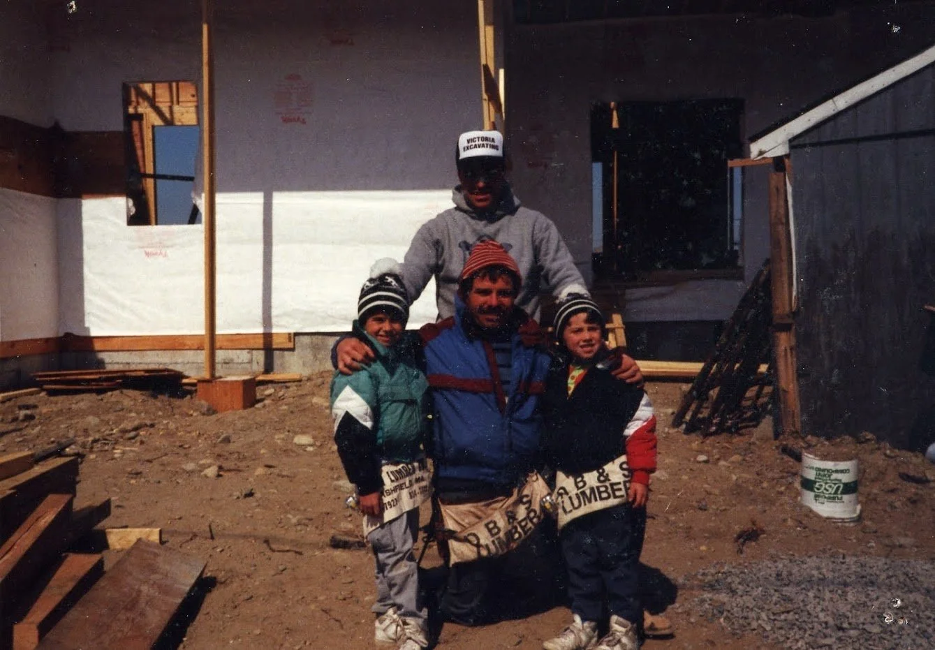Three children and one adult outdoors at a construction site with wooden framing and dirt, all dressed in warm clothing, including jackets, hats, and gloves, during daytime.