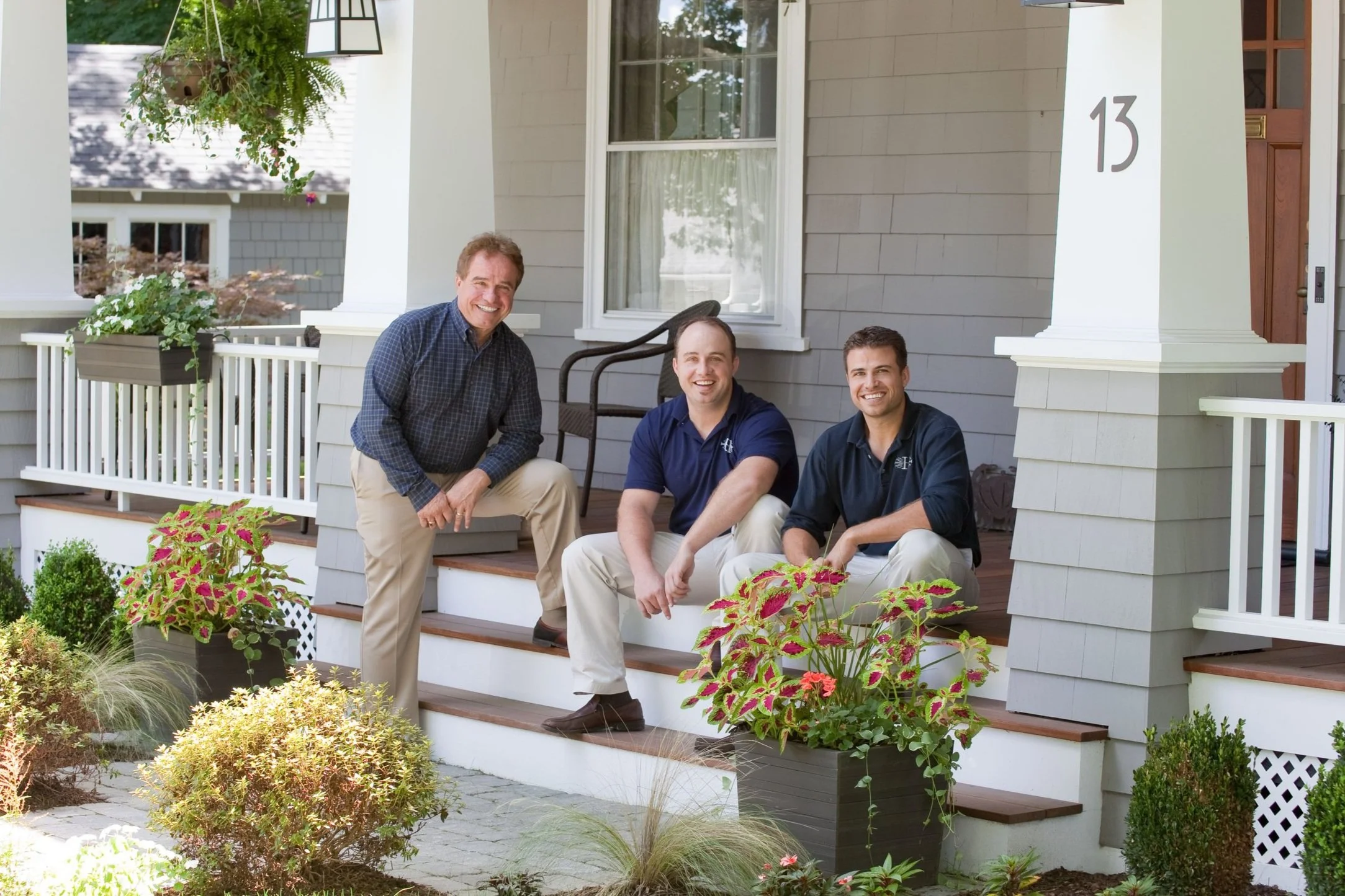 Three men sitting on the front steps of a house, smiling at the camera, surrounded by potted plants and greenery.