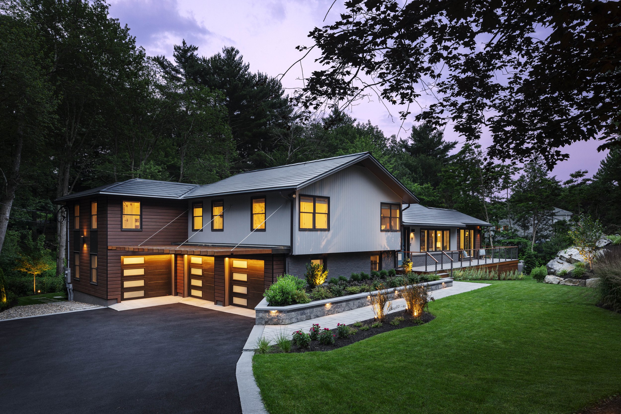 Modern two-story house with a sloped gray roof, a mix of gray and brown exterior walls, a driveway, a landscaped front yard with green grass, bushes, and flowers, and surrounded by trees.