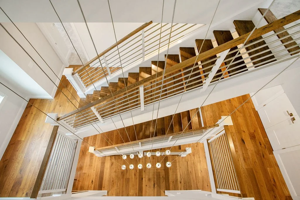 From the top view of a staircase, showing wooden steps, white railing, and a modern chandelier hanging from the ceiling with multiple bulbs.