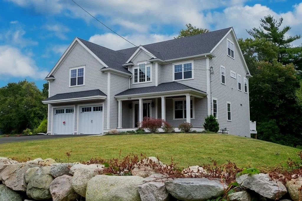 A large two-story house with gray siding, a dark shingle roof, and a attached garage, situated on a grassy lawn with a rock border.